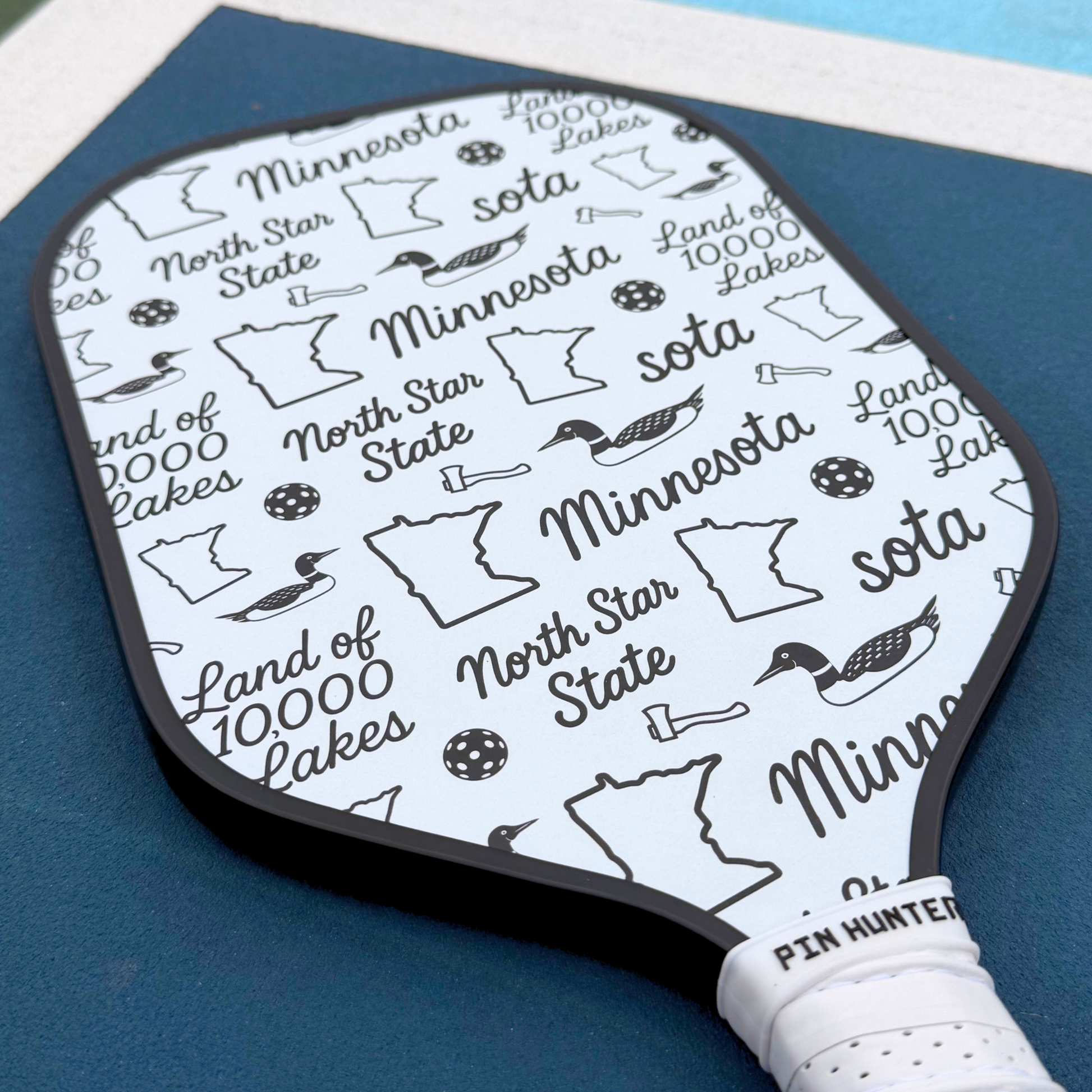 Close-up of Minnesota pickleball paddle with black and white repeating loon, state outline, pickleball, and North Star State icons.