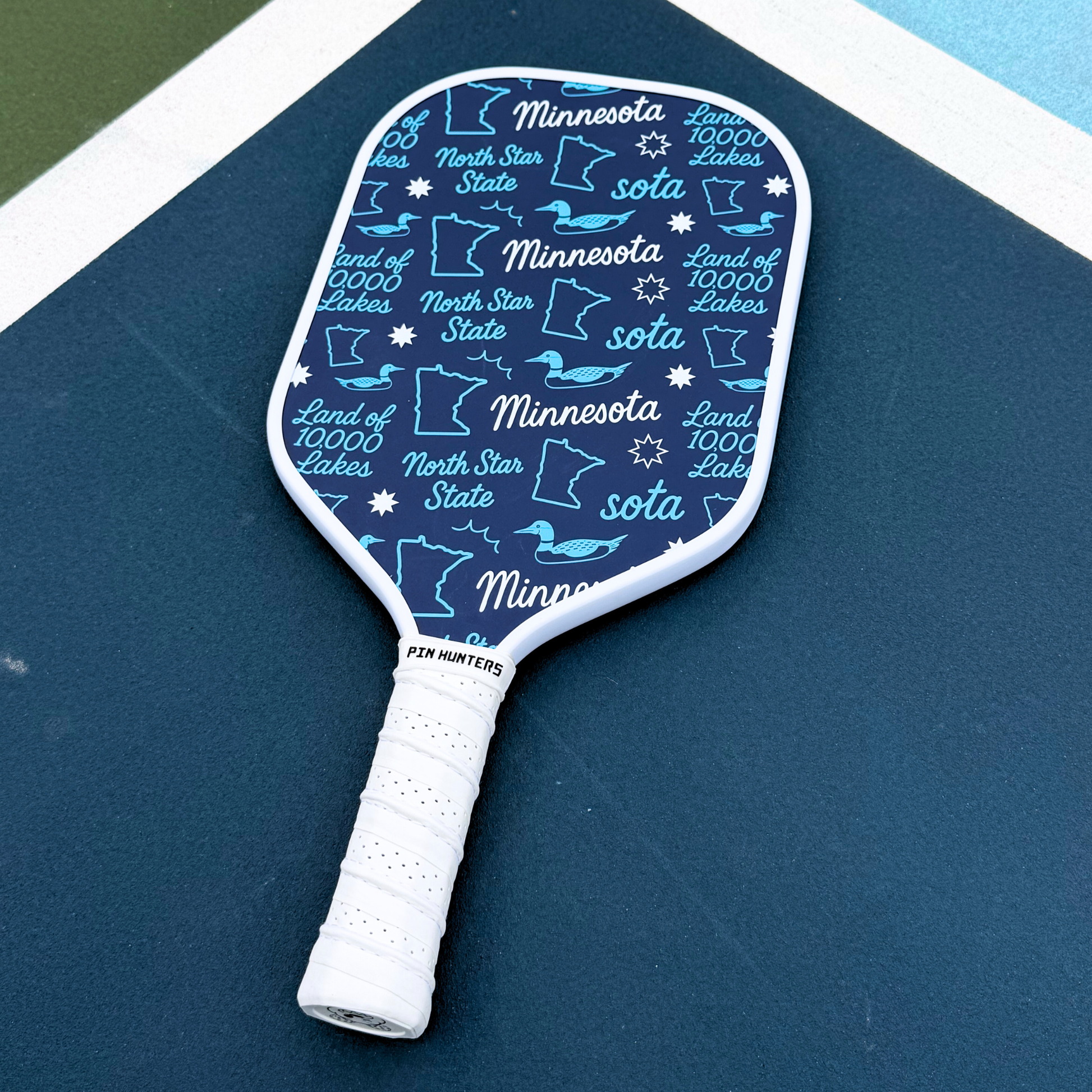 Blue Minnesota pickleball paddle on outdoor court with white trim, white grip, and repeating loon, state outline, and Land of 10,000 Lakes icons.