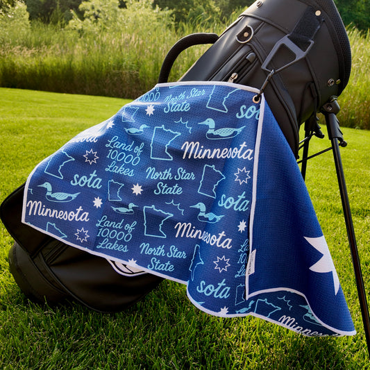 Minnesota flag golf towel hanging on a black golf bag with blue Minnesota back pattern, loons, state outlines, North Star State text, Land of 10000 Lakes text, and golf graphics.