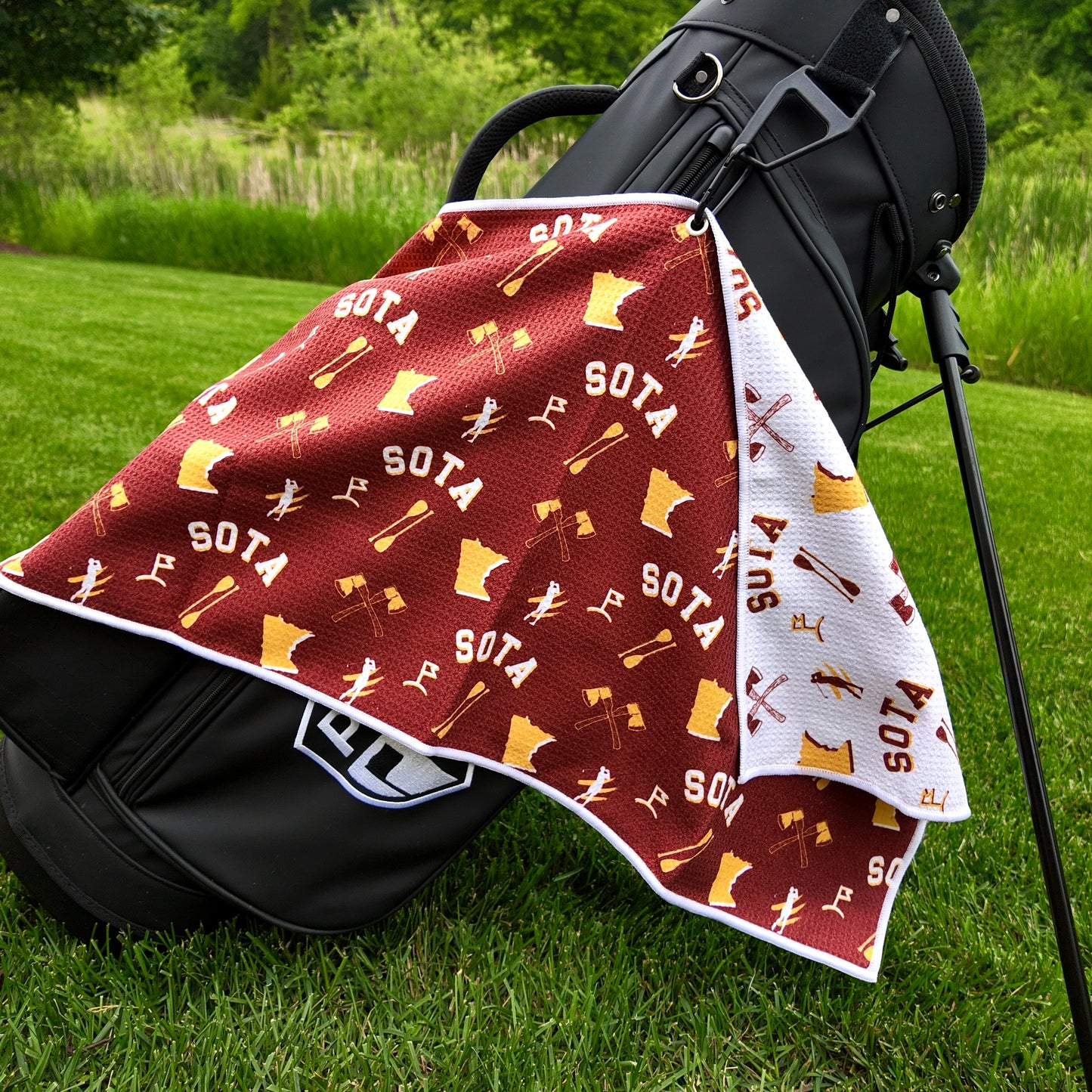 Minnesota Golden Gophers maroon golf towel hanging on a black golf bag with SOTA text, gold Minnesota outlines, crossed oars, golfers, golf flags, and white reverse side.