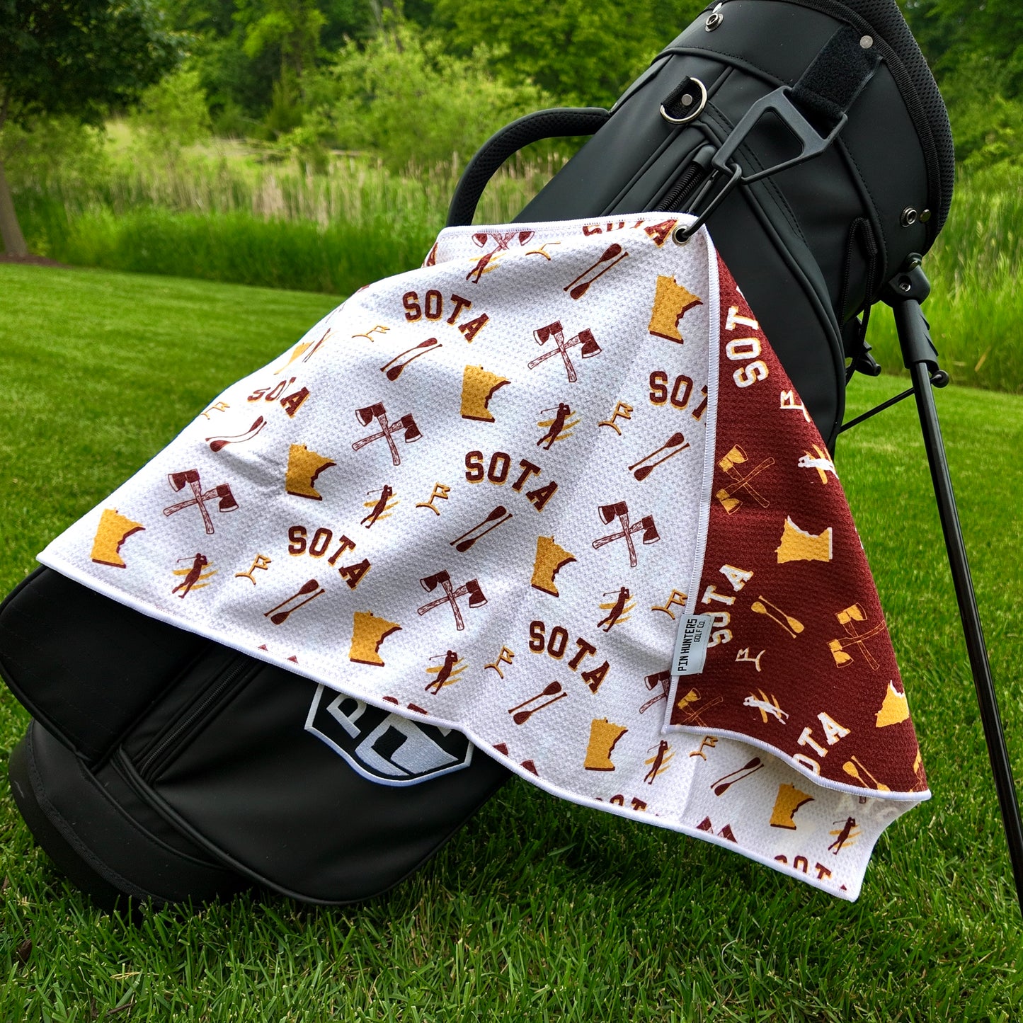 Minnesota Golden Gophers white golf towel hanging on a black golf bag with SOTA text, Minnesota outlines, maroon and gold graphics, crossed oars, golfers, and maroon reverse side.