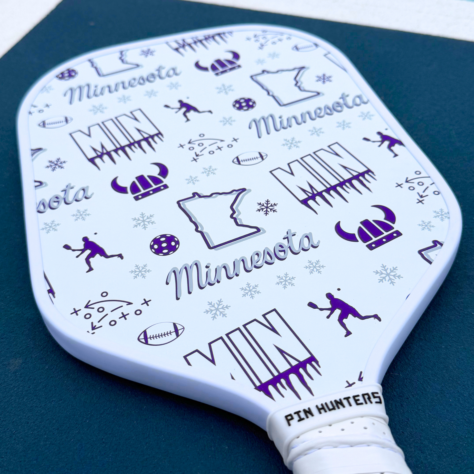 Minnesota Vikings pickleball paddle close-up with white face, purple MIN graphics, football icons, helmet icons, snowflakes, and white handle.