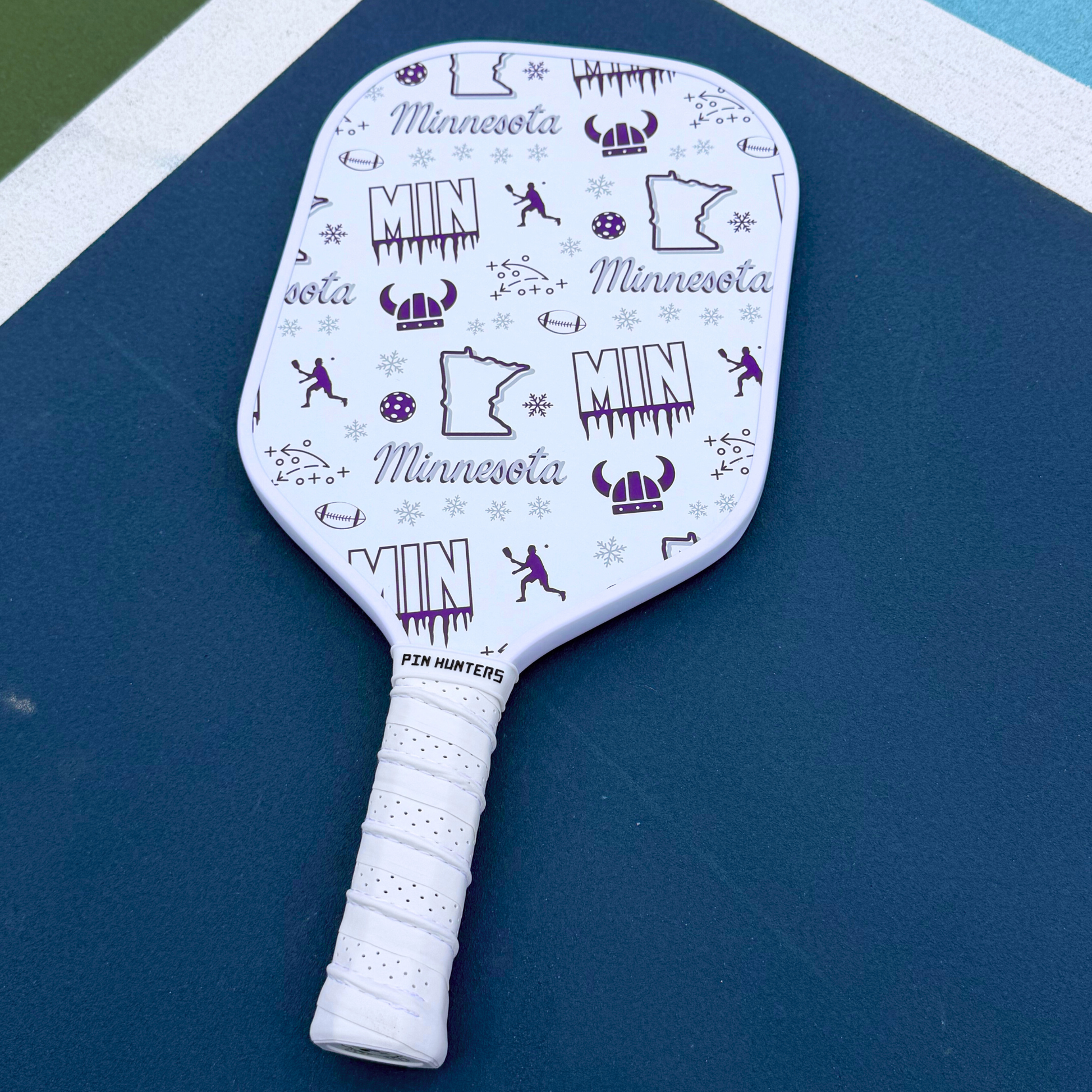 Minnesota Vikings pickleball paddle on court with white face, purple MIN graphics, football icons, snowflakes, helmet icons, and white handle.