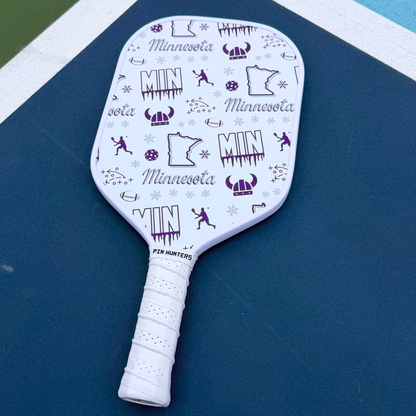 Minnesota Vikings pickleball paddle on court with white face, purple MIN graphics, football icons, snowflakes, helmet icons, and white handle.