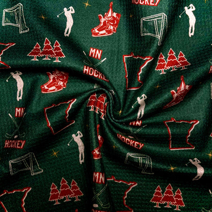 Close up of a green Minnesota Wild inspired hockey golf towel showing MN hockey text, red skates, hockey nets, Minnesota outlines, pine trees, golfers, and waffle microfiber texture.