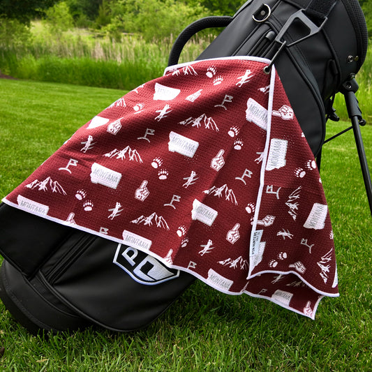 Montana Grizzlies maroon golf towel hanging on a black golf bag with Montana text, mountain graphics, bear paw prints, foam fingers, golfers, and golf flags.