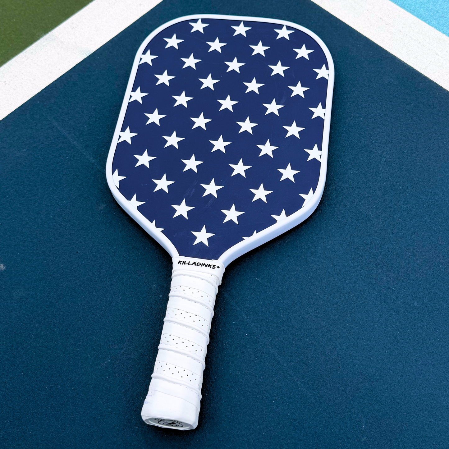 Navy pickleball paddle on court with white star pattern, white edge guard, white grip, and Killa Dinks branding.