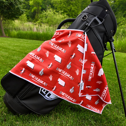 Nebraska Cornhuskers red golf towel hanging on a black golf bag with Nebraska text, state outlines, corn, foam fingers, golfers, and golf icons.