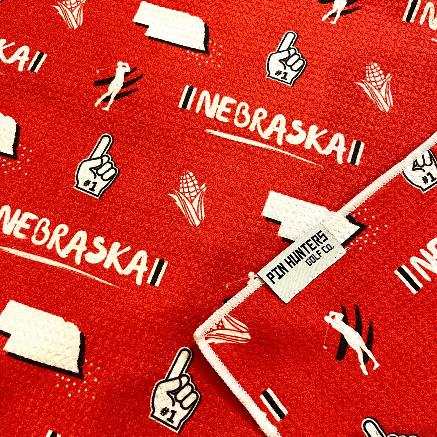 Close up of Nebraska Cornhuskers golf towel with red and white Nebraska, corn, foam finger, state outline, golfer, and golf flag graphics.