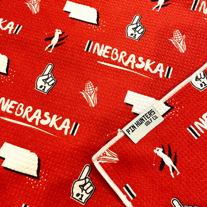 Close up of Nebraska Cornhuskers golf towel with red and white Nebraska, corn, foam finger, state outline, golfer, and golf flag graphics.