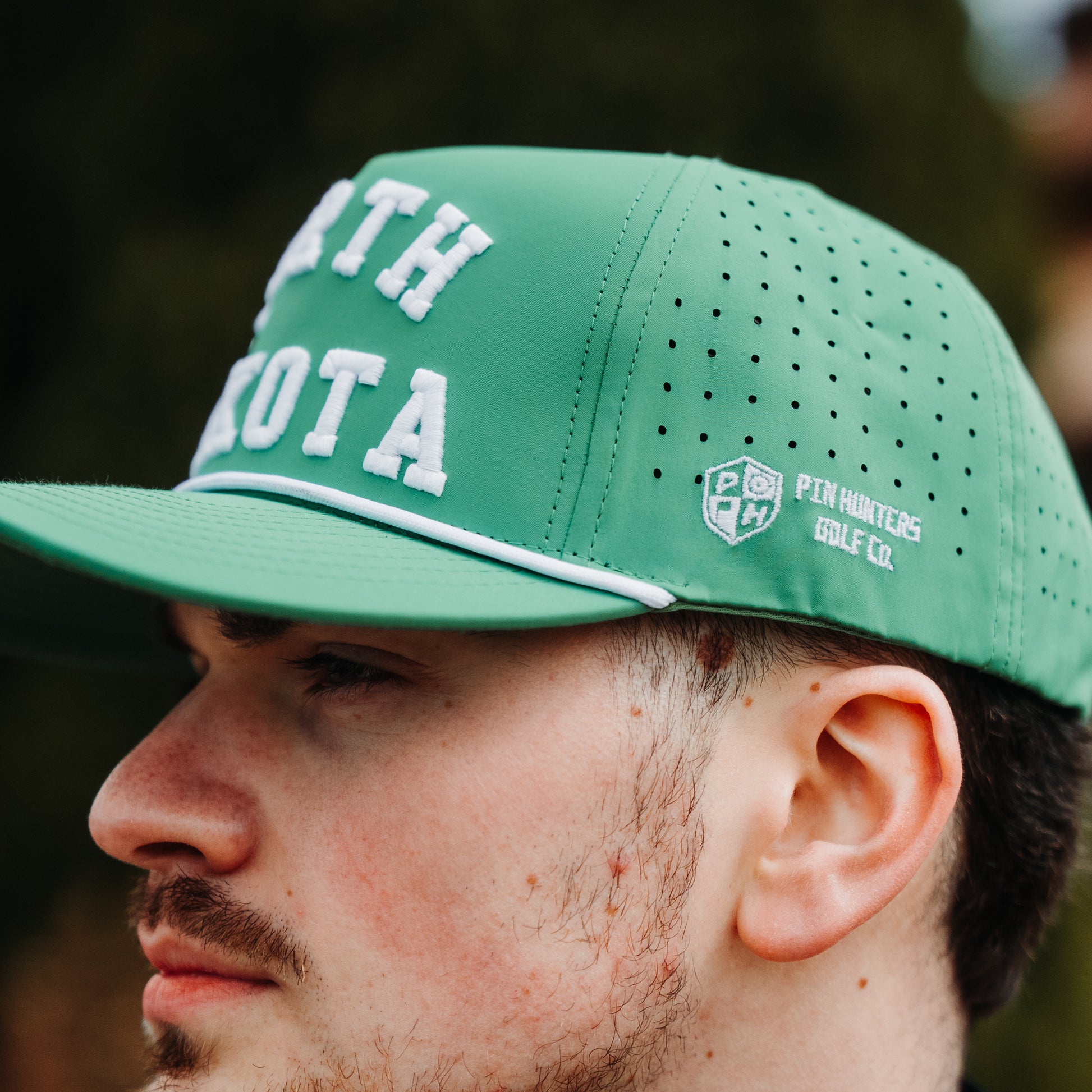 Green North Dakota performance hat lifestyle side view with white embroidery, perforated panels, white rope, and PH logo.