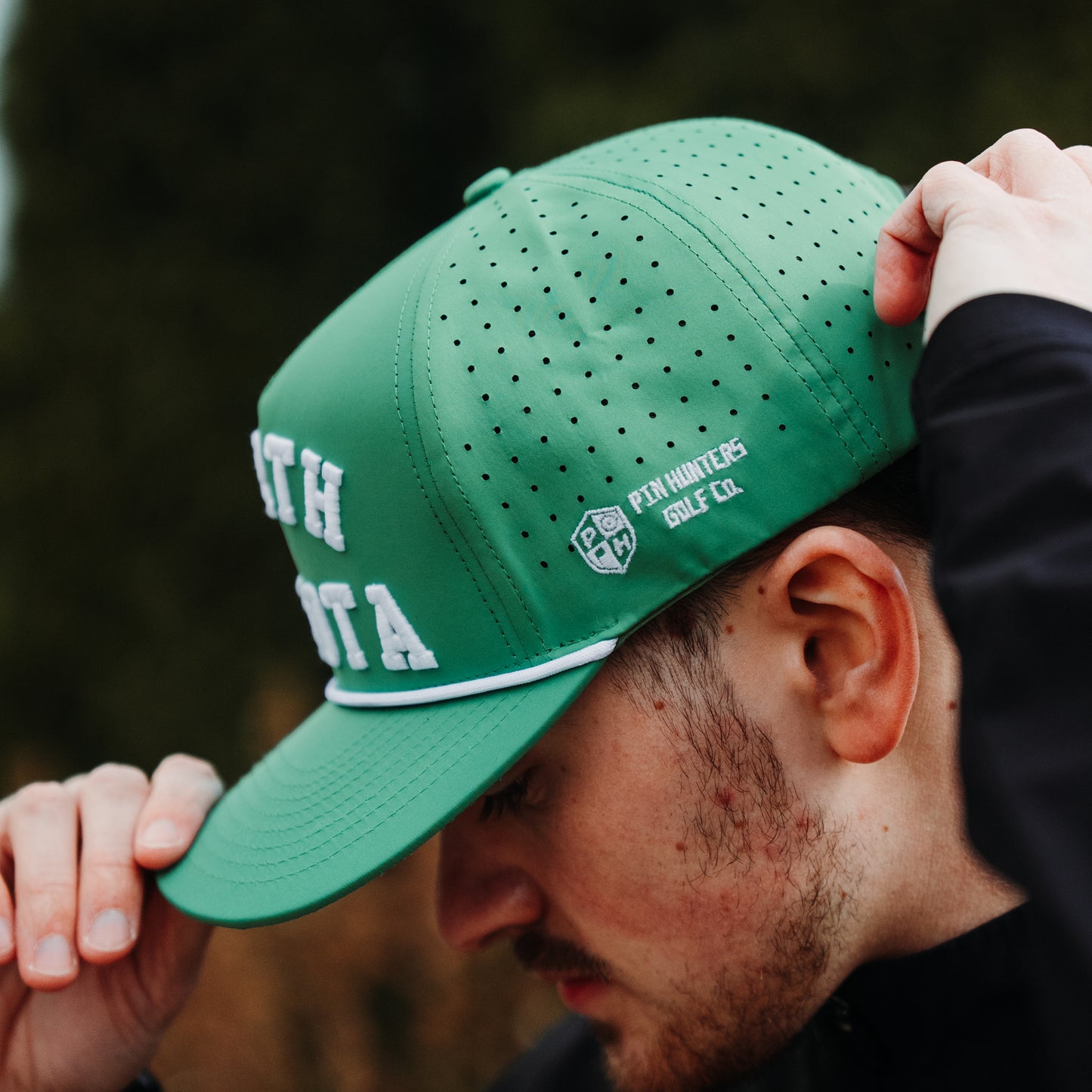 Green North Dakota performance hat side view with white embroidery, white rope detail, perforated side panels, and model wearing the hat.