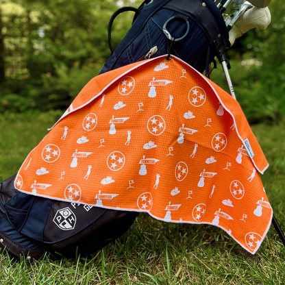 Orange Tennessee TriStar golf towel hanging from a golf bag, showing the orange back pattern with Tennessee TriStar logos, Tennessee text, golf flags, golfers, and golf icons.