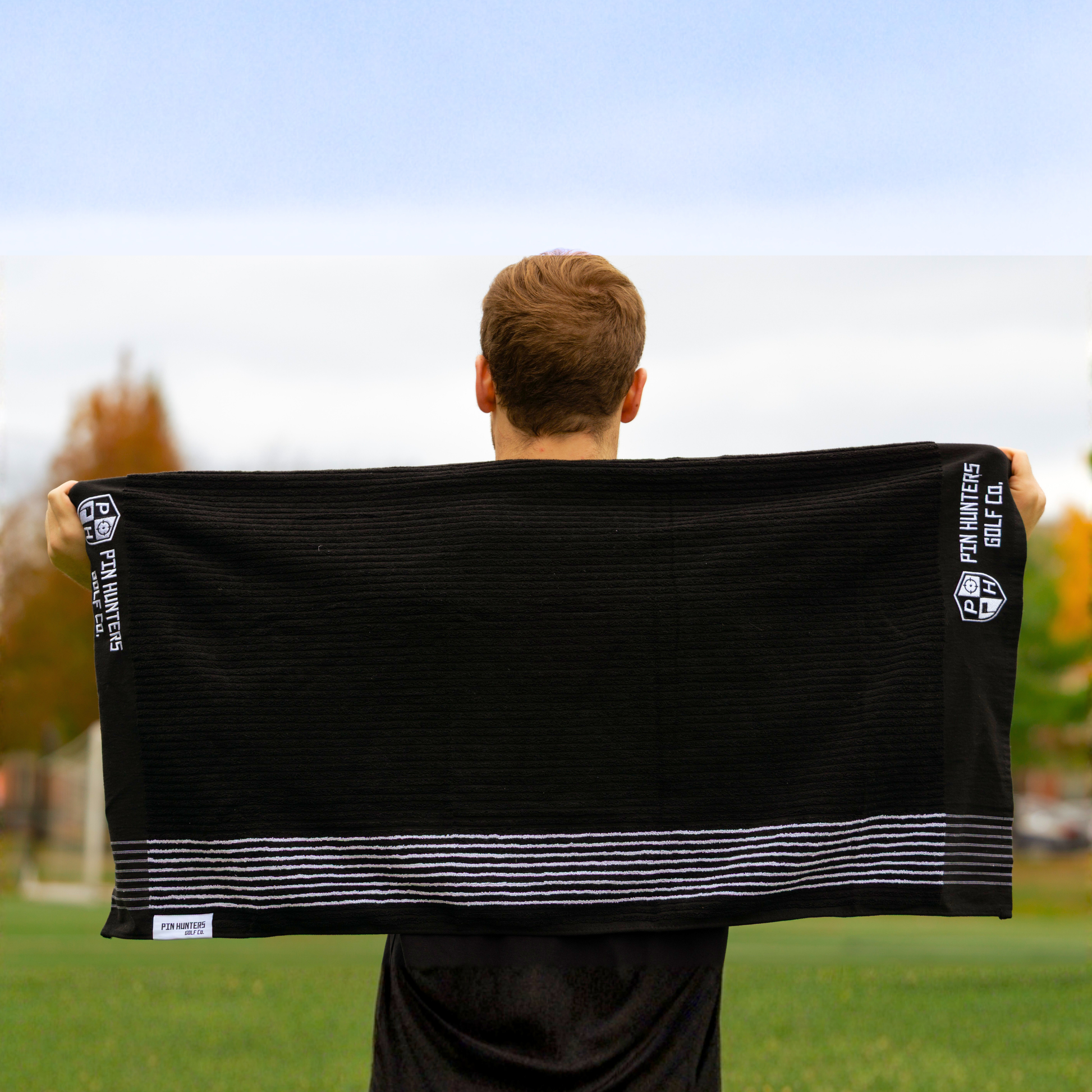 Person holding the Pin Hunters Golf Co. Looper black cotton golf towel fully extended outdoors, showing the full towel shape and white stripe detail.