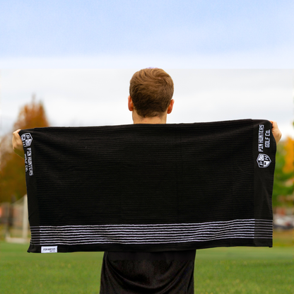 Person holding the Pin Hunters Golf Co. Looper black cotton golf towel fully extended outdoors, showing the full towel shape and white stripe detail.