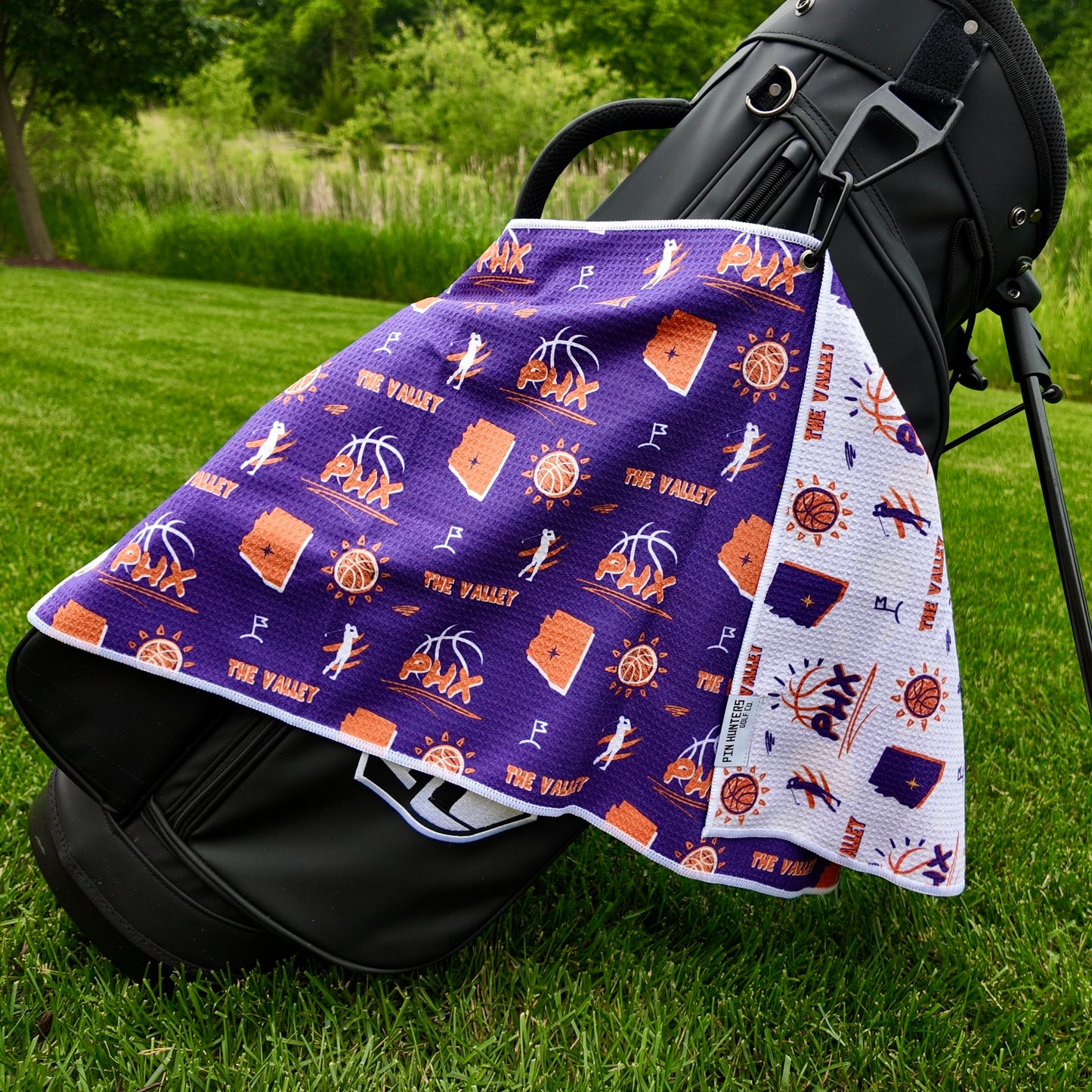 Phoenix Suns purple golf towel hanging on a black golf bag with PHX logos, The Valley text, Arizona outlines, basketball icons, golfers, and white reverse side.