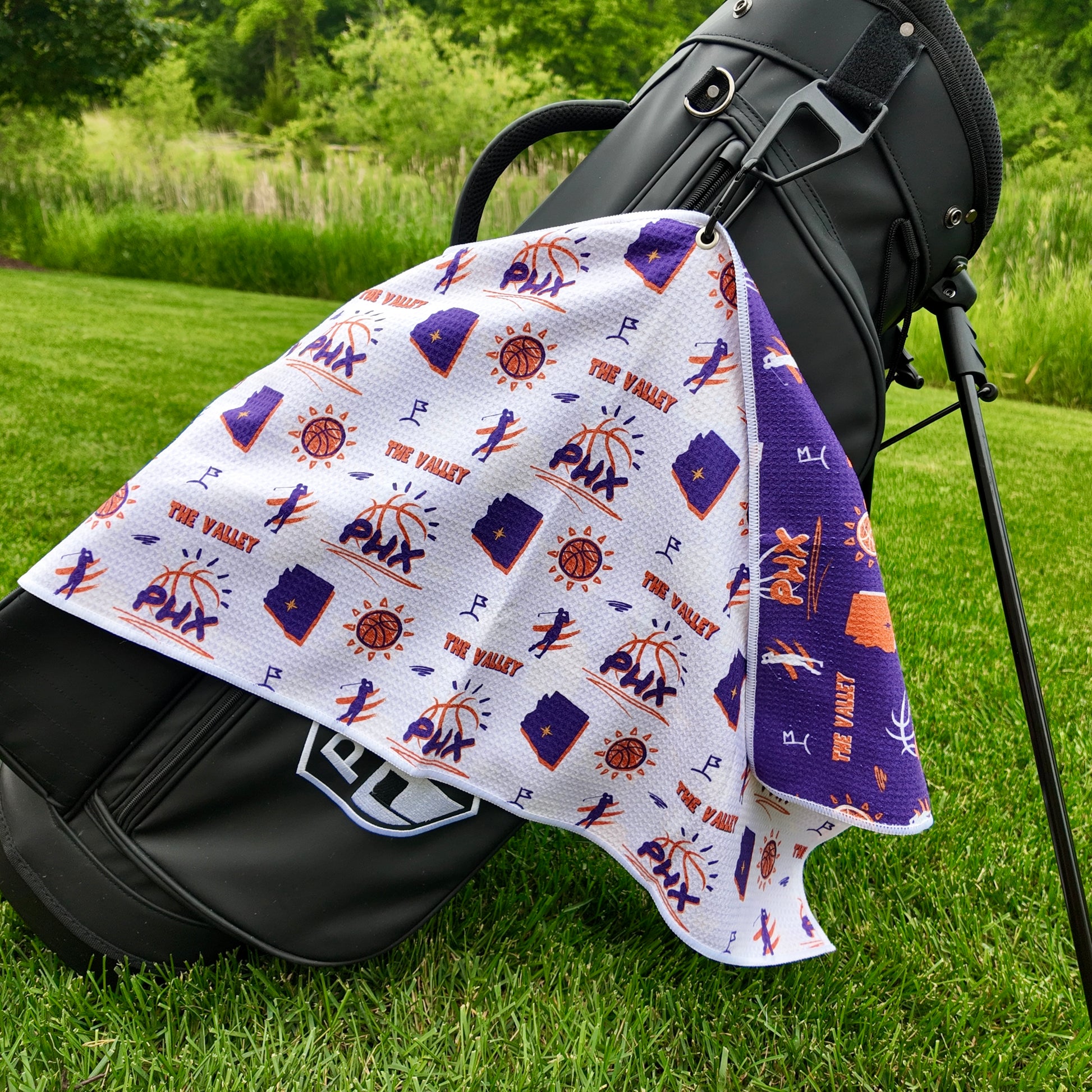 Phoenix Suns white golf towel hanging on a black golf bag with PHX logos, The Valley text, Arizona outlines, basketball icons, golfers, and purple reverse side.