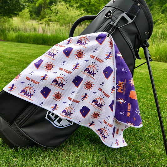 Phoenix Suns white golf towel hanging on a black golf bag with PHX logos, The Valley text, Arizona outlines, basketball icons, golfers, and purple reverse side.