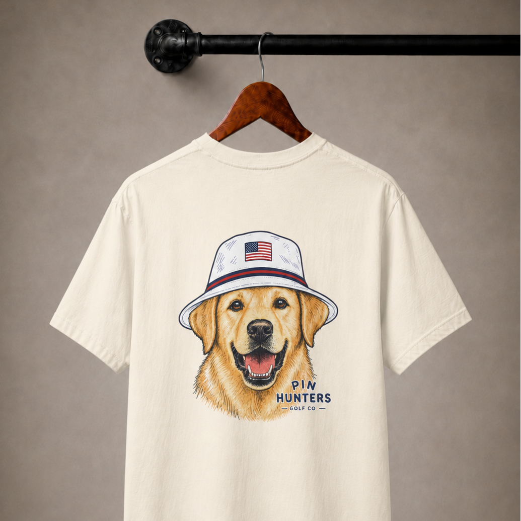 Beige/tan Pin Hunters Colby Tee displayed on a wooden hanger with golden retriever golf graphic and Pin Hunters logo on the back.