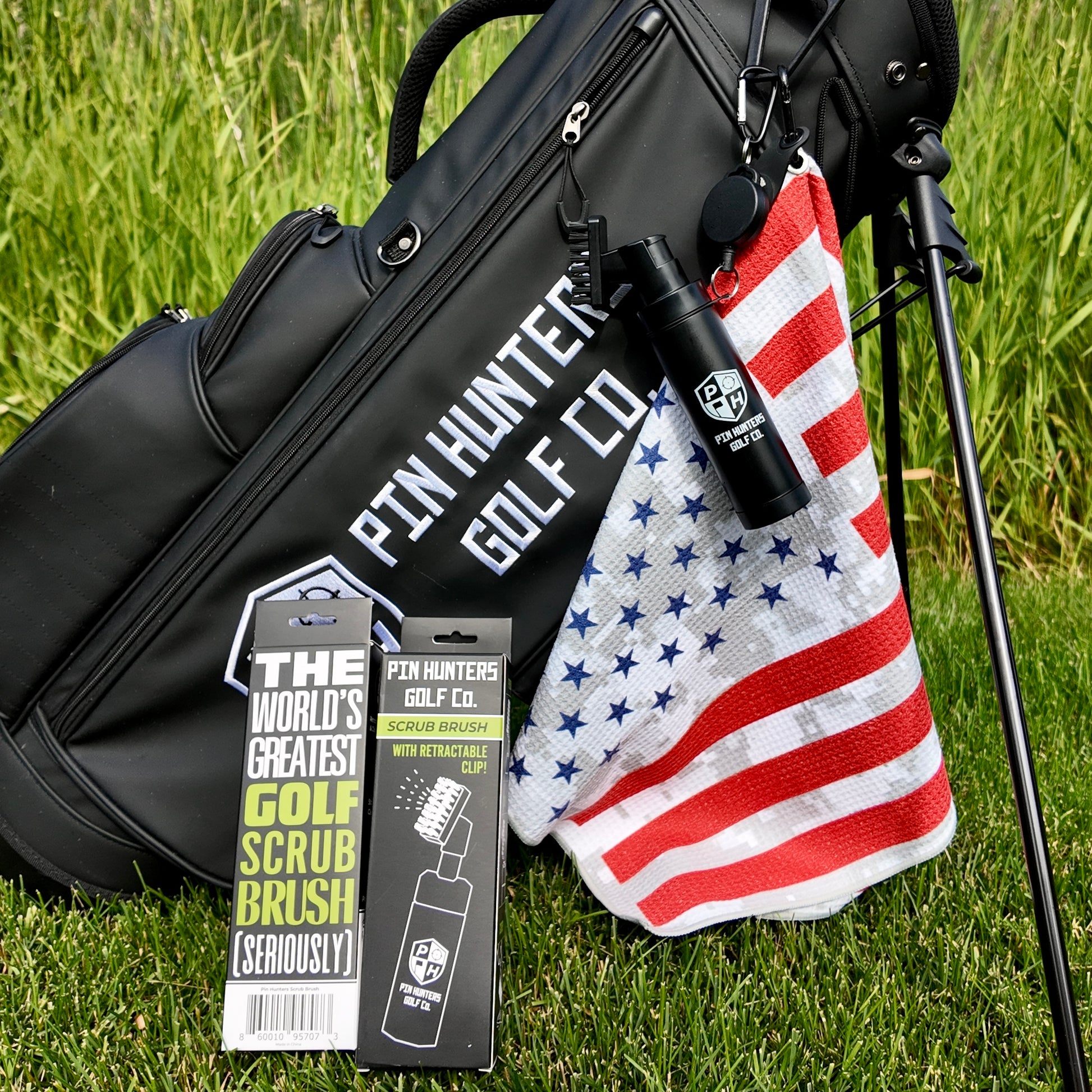 Pin Hunters golf bag with American flag golf towel, black scrub brush clipped to the bag, and scrub brush retail packaging on grass.