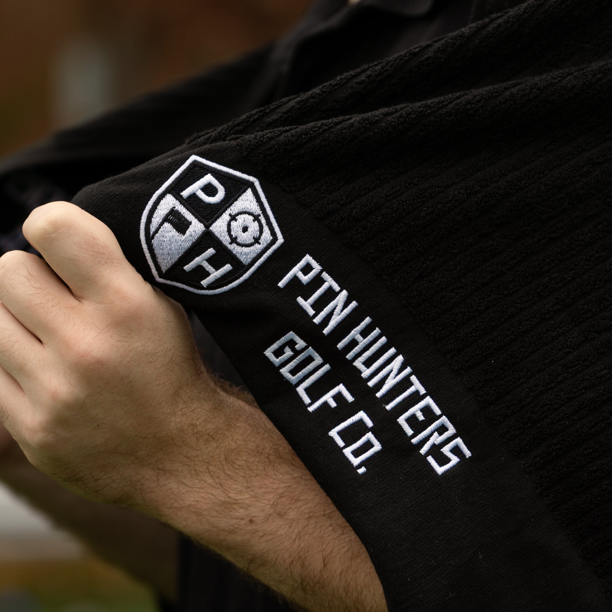 Close-up of the Pin Hunters Golf Co. Looper black cotton golf towel showing the embroidered PH logo and white Pin Hunters Golf Co. branding.