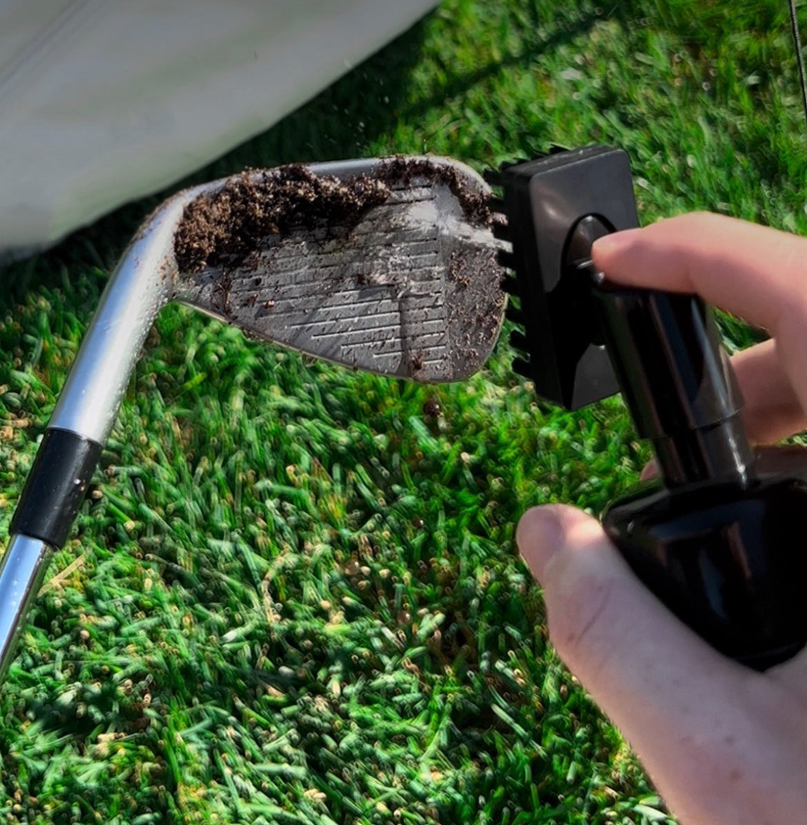 Pin Hunters golf scrub brush spraying water onto a dirty golf club face on turf, showing the brush bottle in use for cleaning grooves.
