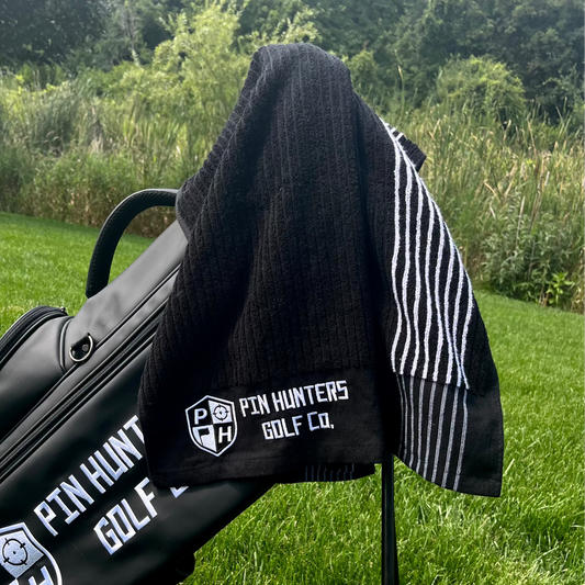 Pin Hunters Golf Co. Looper black cotton golf towel draped over a black golf bag outdoors on the course.