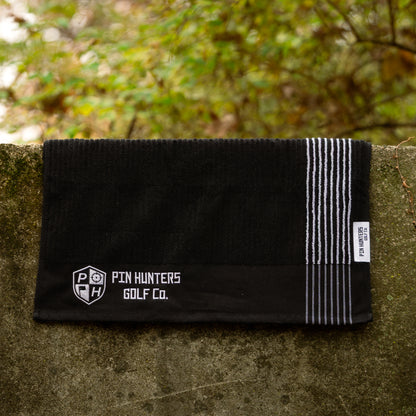 Pin Hunters Golf Co. Looper black cotton golf towel with white stripes hanging on an outdoor stone wall.