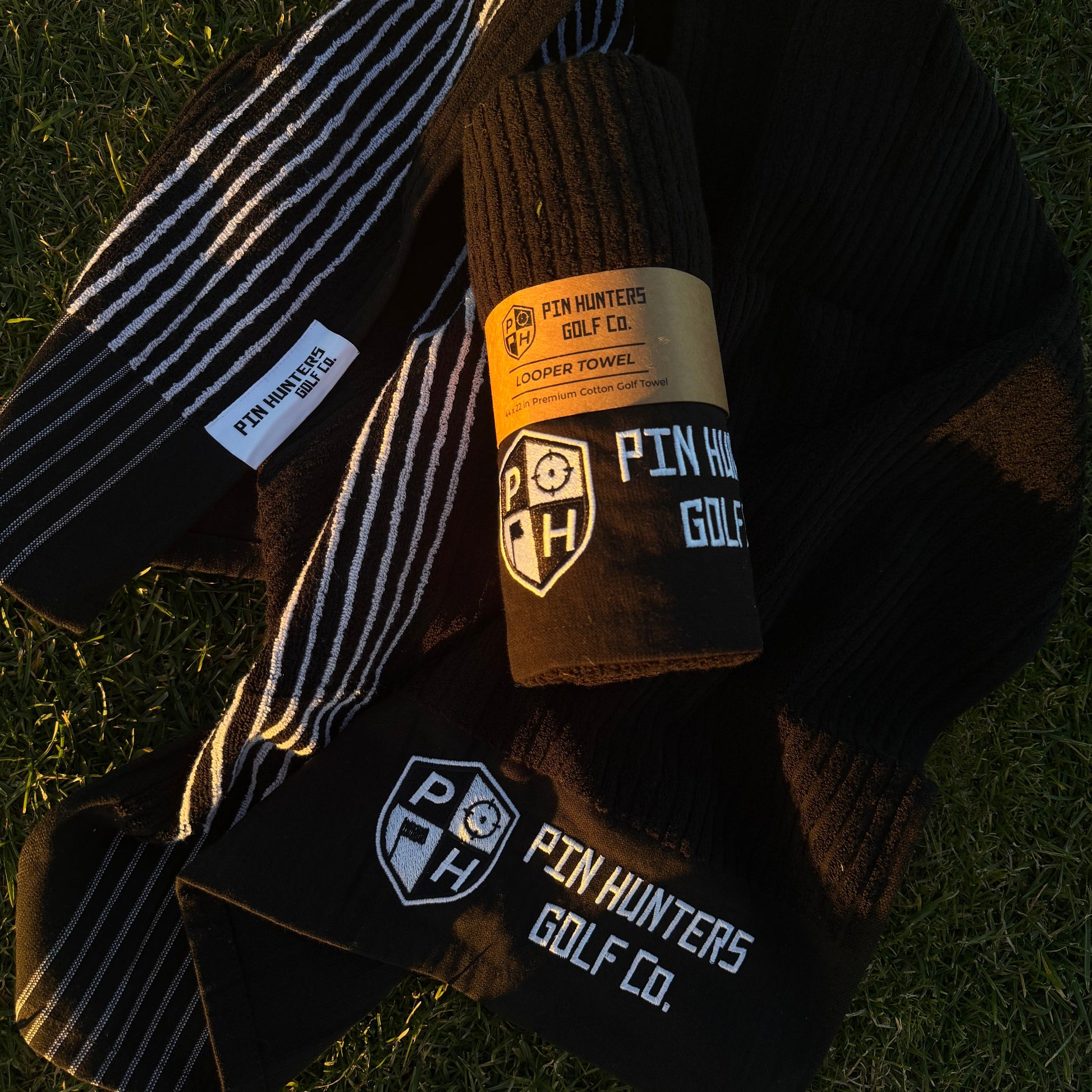 Pin Hunters Golf Co Looper premium cotton golf towel, black knit and black-and-white stripe towels on grass.