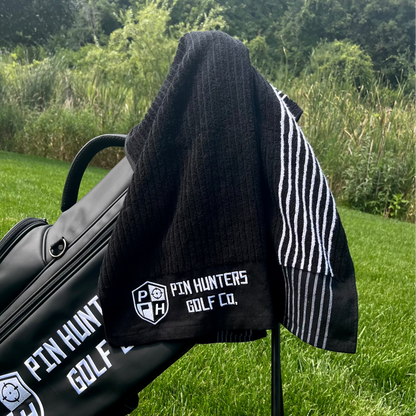 Pin Hunters Golf Co Looper black cotton golf towel with white stripes hanging on a golf bag outdoors on the course.