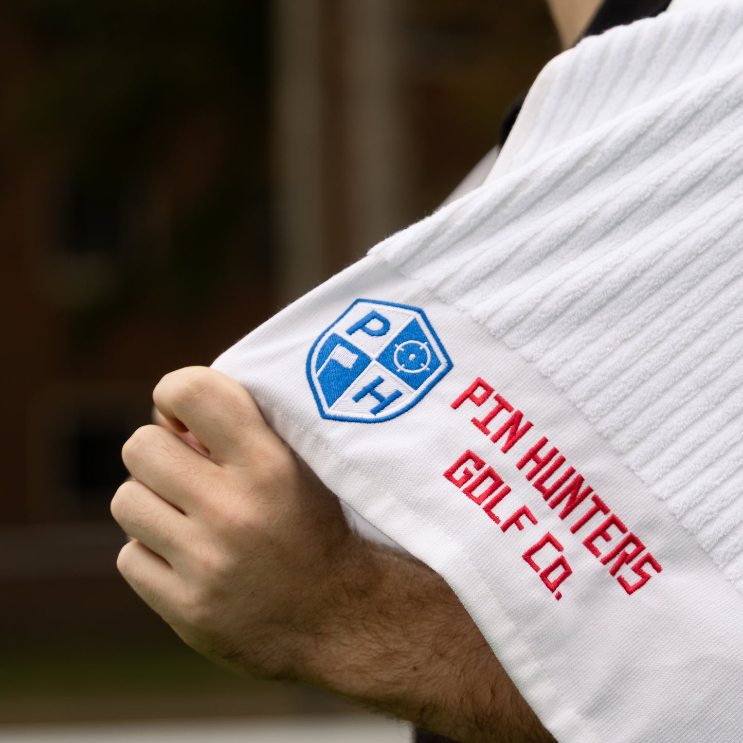 Close up of white Pin Hunters Golf Co. looper towel with blue shield logo, red embroidered wordmark, ribbed cotton texture, and hand holding the towel.