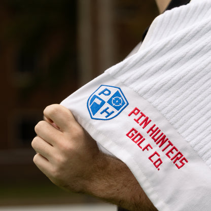 Close up of white Pin Hunters Golf Co. looper towel with blue shield logo, red embroidered wordmark, ribbed cotton texture, and hand holding the towel.