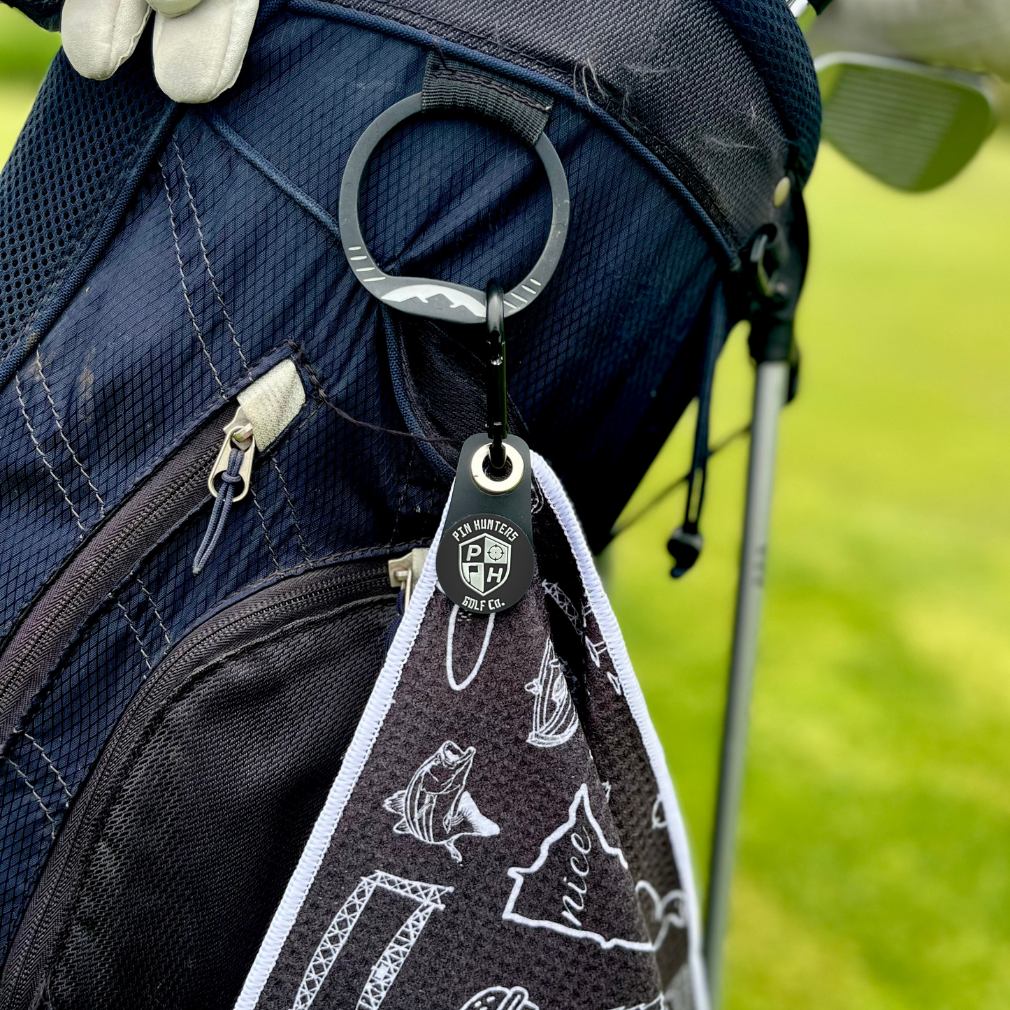 Pin Hunters magnetic towel adapter clipped to a golf towel and attached to a navy golf bag, showing the black PH logo magnet and removable attachment system.