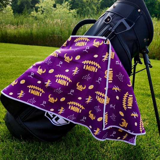 Purple Minnesota Vikings inspired golf towel hanging from a black golf bag with gold football, helmet, horn, MIN, and golf graphics.