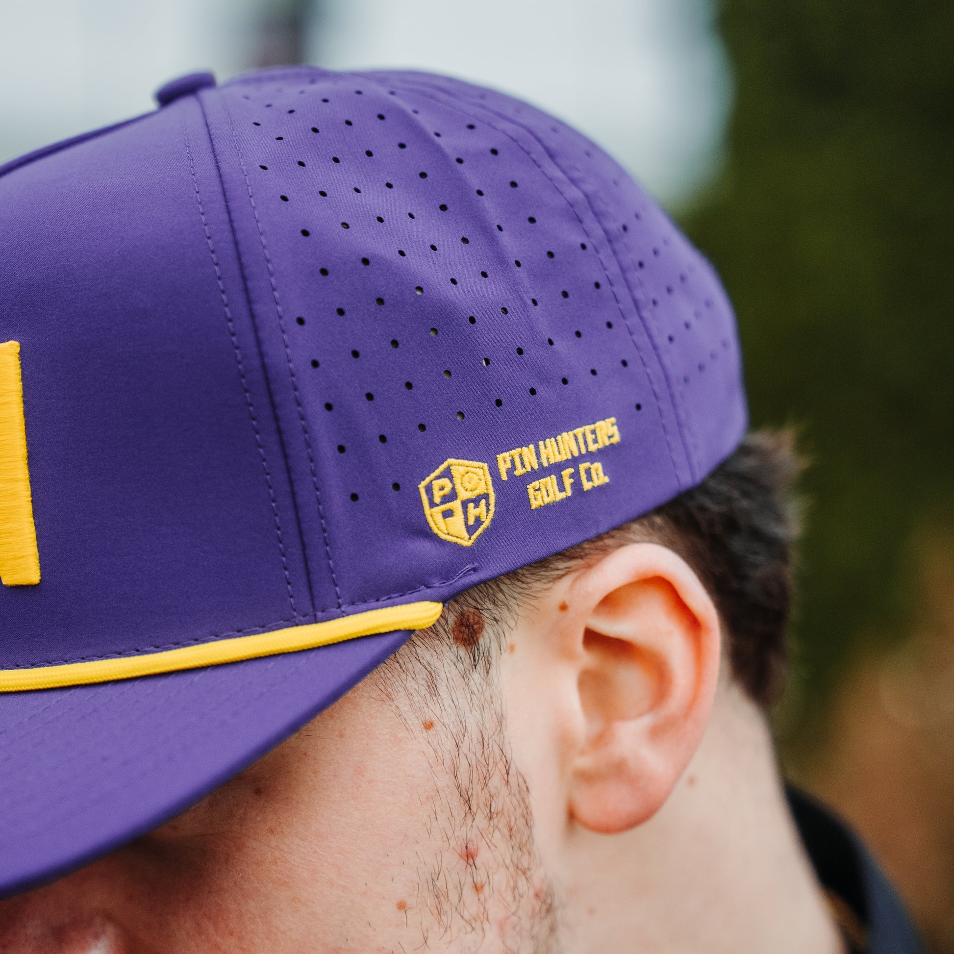 Close up of purple performance hat with yellow Pin Hunters Golf Co. side embroidery, perforated side panels, and yellow rope.