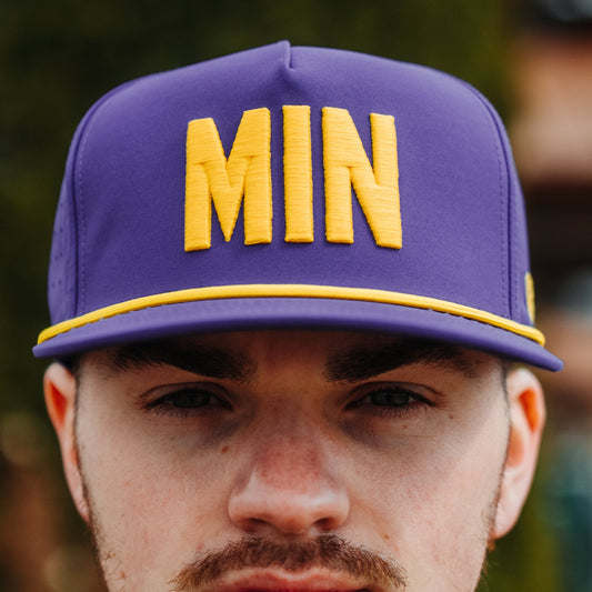 Purple MIN performance golf hat with yellow raised embroidery, yellow rope detail, perforated side panels, and model front view.