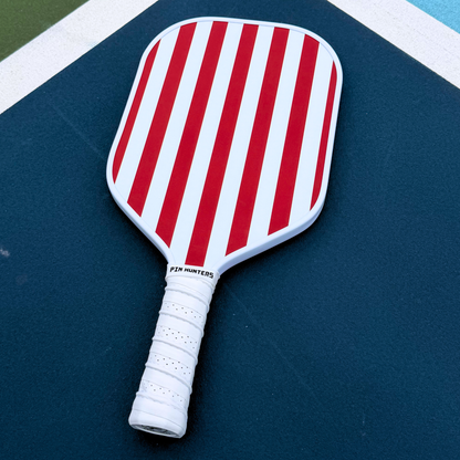 USA themed pickleball paddle on court with red and white vertical stripe design, white edge guard, and white grip.