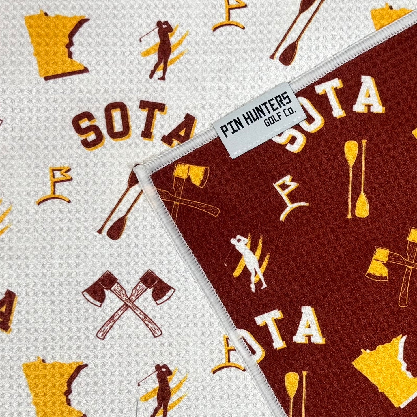 Close up of SOTA golf towel with maroon and gold Minnesota icons, crossed axes, state outlines, golf graphics, and Pin Hunters Golf Co. label.