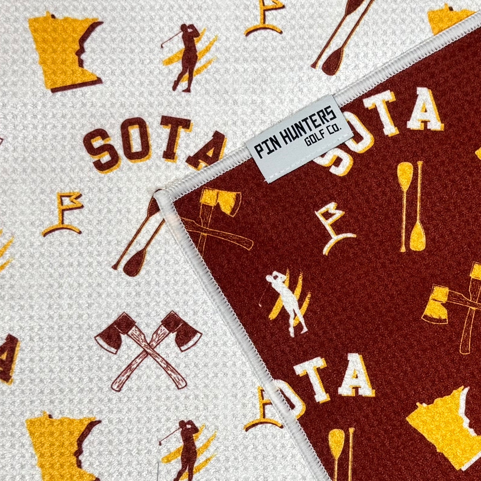 Close up of SOTA golf towel with maroon and gold Minnesota icons, crossed axes, state outlines, golf graphics, and Pin Hunters Golf Co. label.