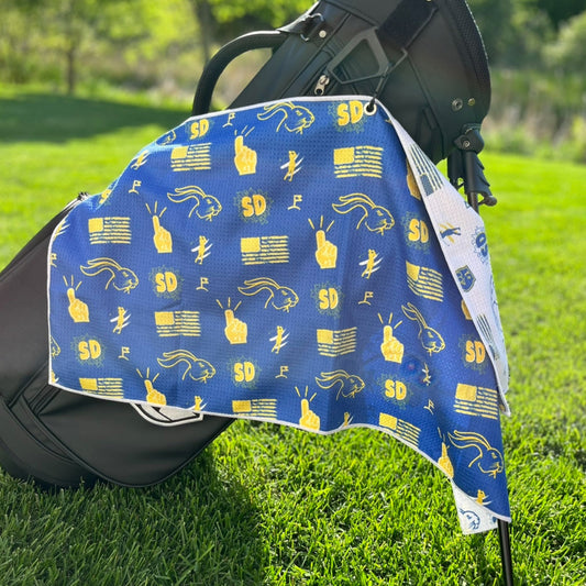 South Dakota State golf towel hanging on a golf bag with blue and yellow SD, jackrabbit, football, golfer, and flag graphics.
