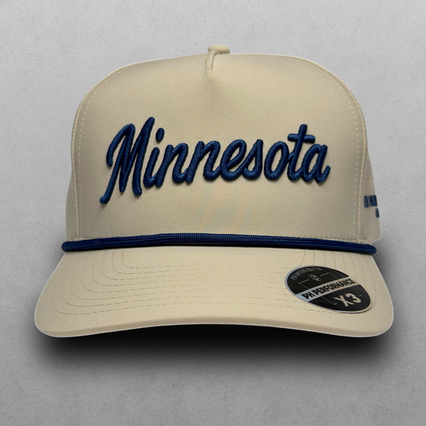 Studio front view of tan Minnesota script performance golf hat with navy embroidery, navy rope, flat brim, and PH Performance X3 sticker.
