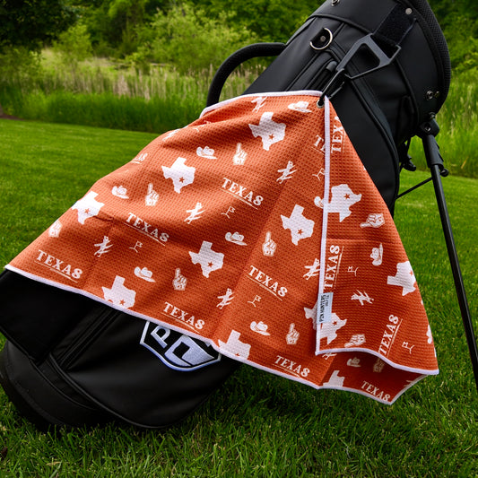 Texas Longhorns burnt orange golf towel hanging on a black golf bag with Texas text, white Texas outlines, foam fingers, golfers, golf flags, and white reverse side.