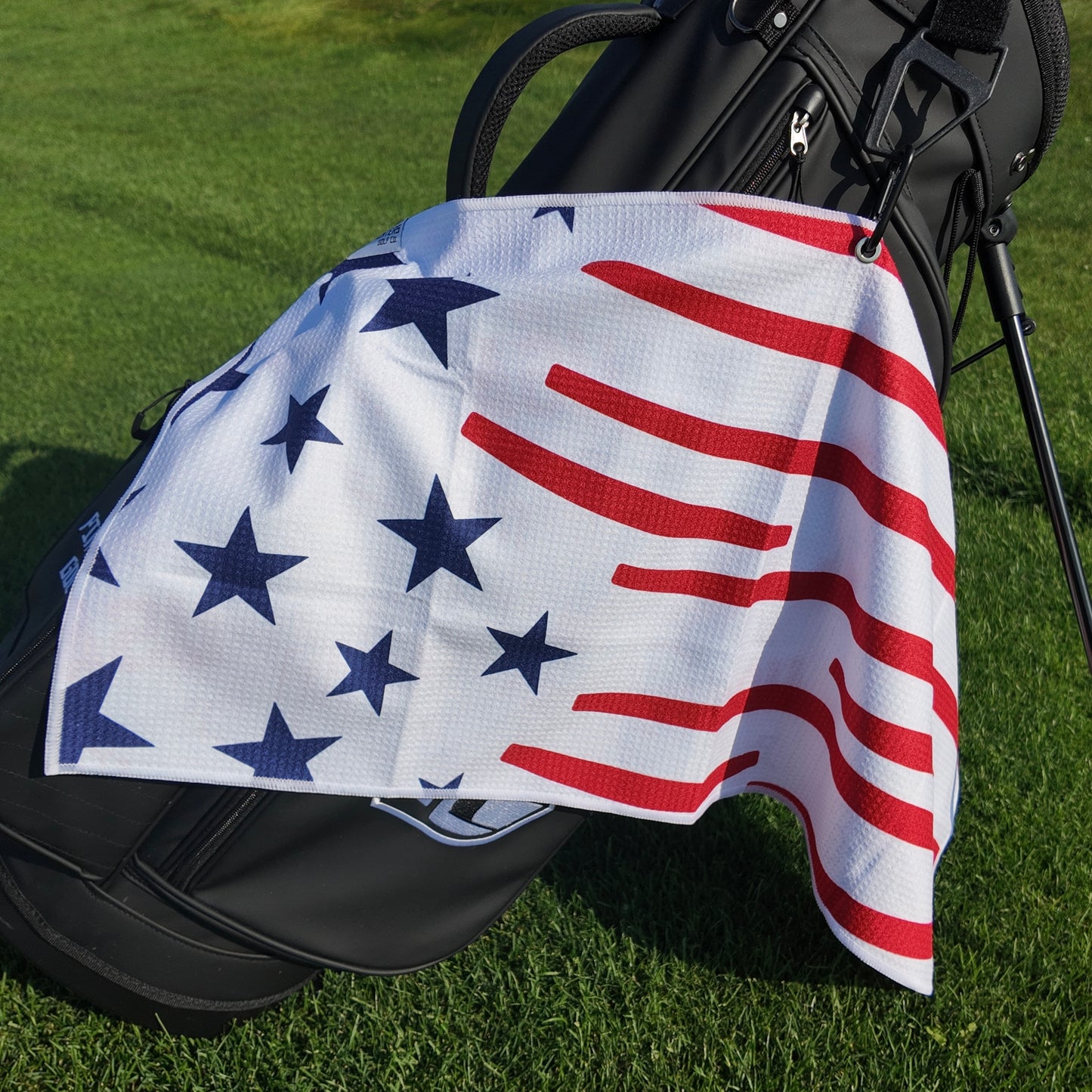 USA 1776 golf towel hanging on a black golf bag with navy stars, red stripes, white microfiber waffle fabric, and patriotic American flag design.