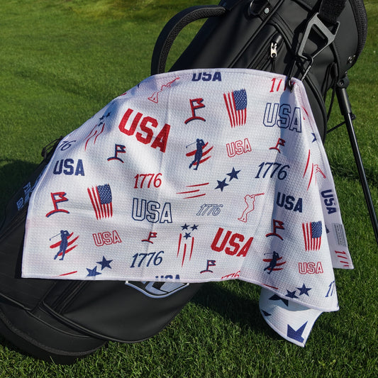 USA 1776 patriotic golf towel hanging on a black golf bag with red, white, and blue USA text, 1776 graphics, American flags, golfers, and golf icons.