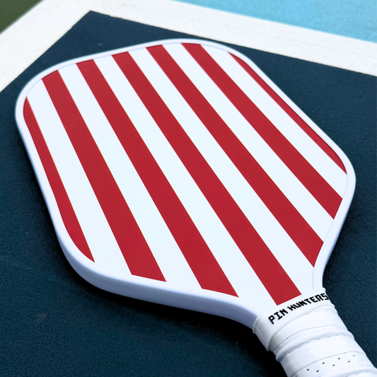 USA themed pickleball paddle on court with red and white vertical stripe design, white edge guard, and white grip.