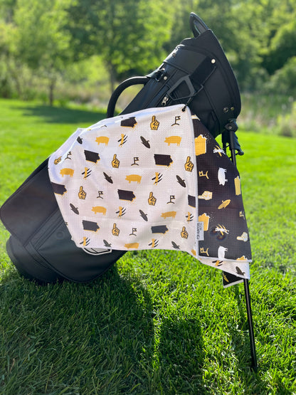 White, black, and gold Iowa Hawkeyes inspired golf towel hanging from a black golf bag with state, corn, pig, foam finger, and golf icons.