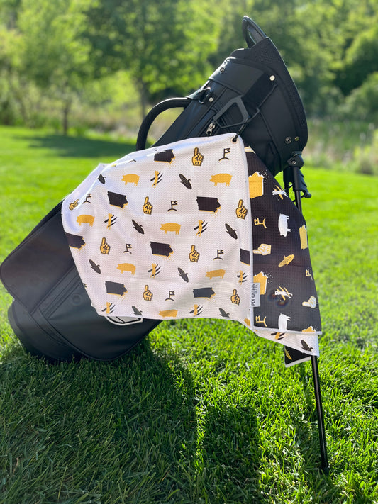 White, black, and gold Iowa Hawkeyes inspired golf towel hanging from a black golf bag with state, corn, pig, foam finger, and golf icons.