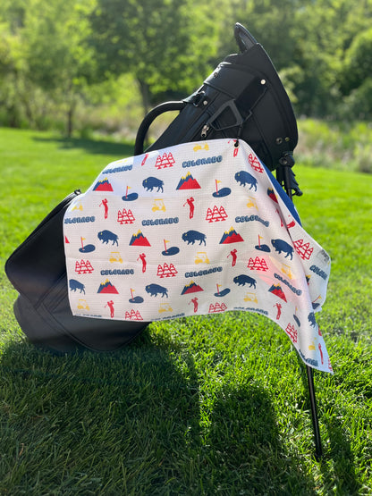 White Colorado golf towel hanging from a black golf bag with colorful Colorado icons, including mountains, bison, pine trees, golfers, golf carts, and flags.