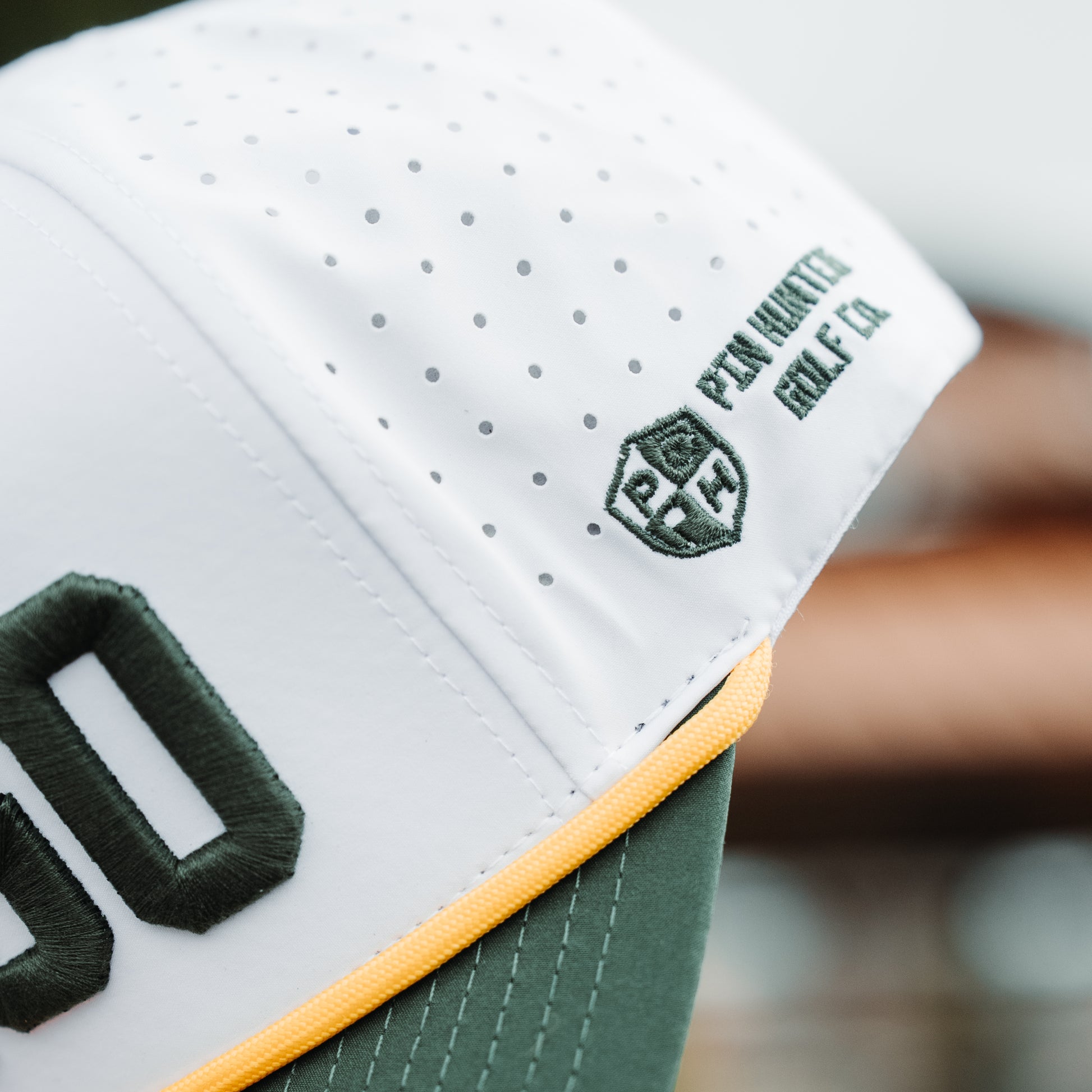 White GB performance golf hat side detail with green Pin Hunters logo embroidery, yellow rope detail, green brim, and perforated side panels.