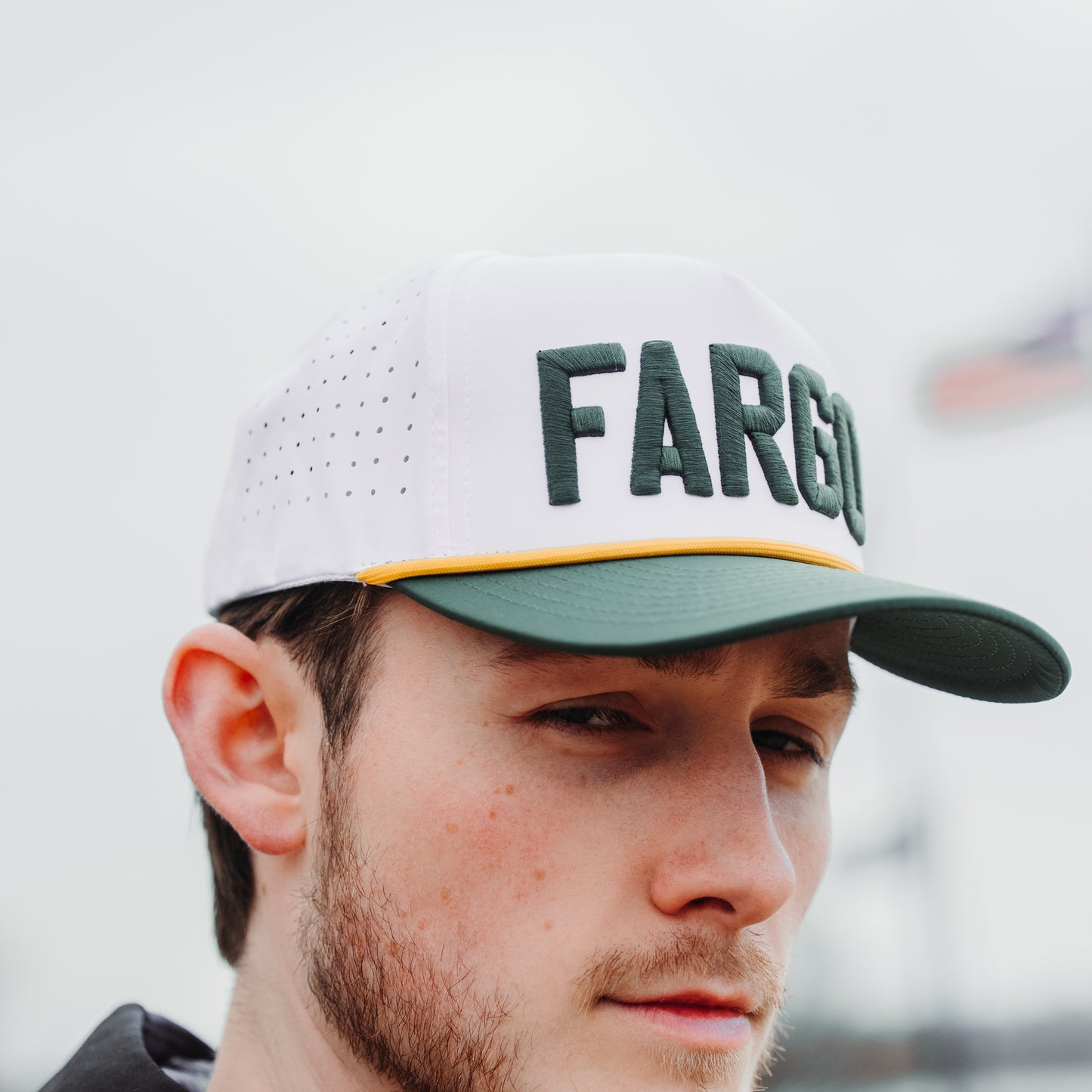 White and green Bay performance golf hat on model with green raised embroidery, green brim, yellow rope detail, and perforated side panels.